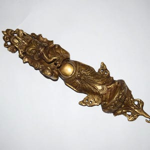 May include: Ornate, golden-colored decorative object with detailed carvings. The design features a figure with an elephant head and multiple arms. The object has a hinged section and is likely a decorative door pull or similar item.