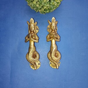 Puede incluir: Dos tiradores de puerta decorativos de color dorado con forma de mujer con cola de serpiente. La mujer está en posición de oración con las manos juntas frente a su pecho.