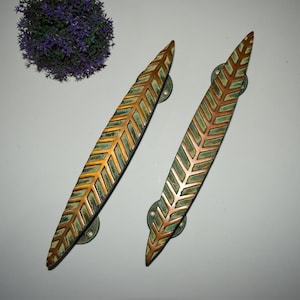 以下が含まれることがあります： 葉のデザインが施された装飾的なドアハンドル2つ。ハンドルは長く細く、銅色の仕上げと緑とクリーム色の模様があります。円形の緑色のベースに取り付けられています。ハンドルの長さは約30cmです。