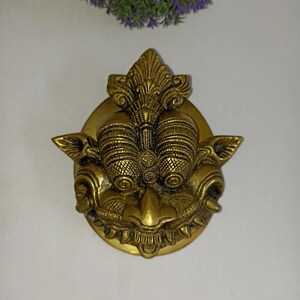 May include: A detailed brass door knocker in the shape of a mythical creature's face. The knocker features intricate carvings, including large eyes, a detailed nose, and a decorative crown. The knocker is mounted on a round brass plate.