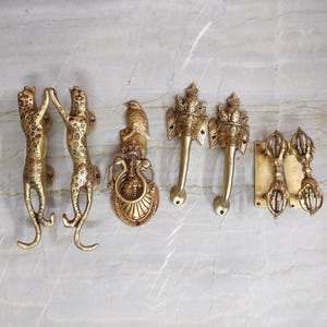 May include: A collection of six brass door handles and knockers. Two handles are shaped like leopards, one is a bird-topped door knocker, two are elephant-headed handles, and one is a double-ended vajra-style handle mounted on a rectangular plate.