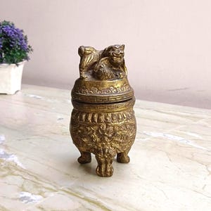 May include: A small, antique brass incense burner with a detailed design. The burner features a lid with a sculpted lion figure and ornate patterns on the body. It stands on three small feet and is displayed on a marble surface.