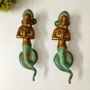 May include: Two bronze-toned mermaid sculptures with green-patina snake tails. The mermaids have their hands clasped in prayer. The sculptures are mounted on a white surface. Decorative art.