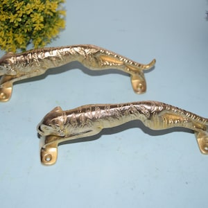 Könnte beinhalten: Zwei goldfarbene Türklinken in Form von springenden Tigern. Die Griffe sind mit strukturiertem Fell verziert und haben eine einfache, rechteckige Basis.