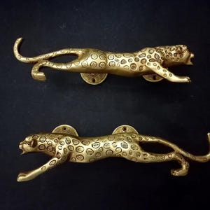 Könnte beinhalten: Zwei Türgriffe aus Messing in Form eines Leoparden. Die Griffe befinden sich in einer Laufposition mit detaillierten Flecken und einer kreisförmigen Montageplatte. Die Griffe sind goldfarben und vor einem dunklen Hintergrund.