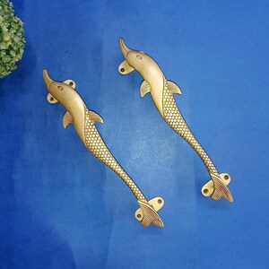 以下が含まれることがあります： 真鍮製のイルカ型ドアハンドル2つ。 ハンドルは金色で、尾とひれにテクスチャがあります。 イルカの体は滑らかで、ハンドルは青い表面に取り付けられています。