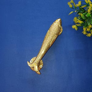 May include: A brass fish-shaped door handle with detailed scale patterns. The handle is mounted on a blue surface, with a small floral arrangement in the upper right corner. The fish handle is approximately 25 cm long.