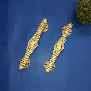 May include: Two brass door handles with floral and geometric patterns. The handles have a long, rectangular shape with decorative detailing and circular mounting plates. The handles are set against a blue background.