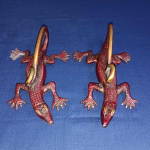 Könnte beinhalten: Zwei dekorative Eidechsenfiguren. Die Eidechsen sind rotbraun mit goldfarbenen Schwänzen und Akzenten. Die Eidechsen liegen auf einer blauen Oberfläche. Die Eidechsen haben detaillierte Schuppen und Krallenfüße.
