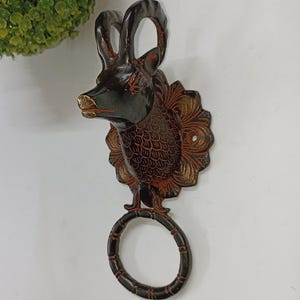 May include: A wall-mounted towel ring with a deer head design. The deer head is dark brown with gold accents on the nose and antlers. The towel ring is a dark brown circular shape. The decorative piece is mounted on a white wall.