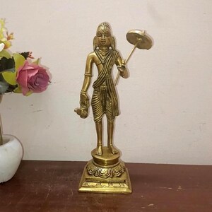 May include: A gold-colored statue of a standing figure holding an umbrella and a small object. The statue is on a square base with ornate detailing. The figure is wearing a draped garment and jewelry.