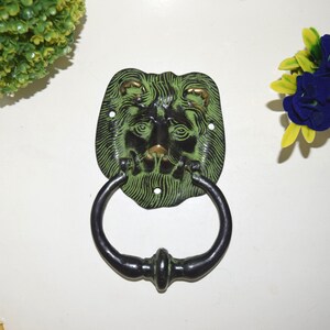 Puede incluir: Un llamador de puerta de cabeza de león verde y negro con un anillo de metal negro. La cara del león está detallada con acentos dorados.