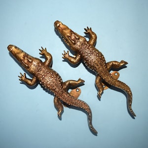 Peut inclure: Deux poignées de porte en forme de crocodile dorées avec des écailles détaillées et des gueules ouvertes. Les poignées sont montées sur un fond bleu clair.