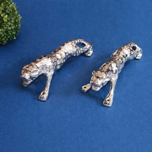 May include: Two silver-colored, decorative cheetah figurines. The cheetahs are in a crawling pose, with detailed spots and textured surfaces. The figurines are set against a blue background, suggesting a decorative or home decor theme.