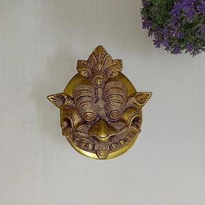 Könnte beinhalten: Ein detailliertes, goldfarbenes Wandbild mit einem stilisierten Gesichtsdesign. Das Gesicht hat große, strukturierte Augen, eine markante Nase und einen detaillierten Mund. Das Stück ist rund und hat ein verziertes kronenartiges Element oben. Der Hintergrund ist weiß.
