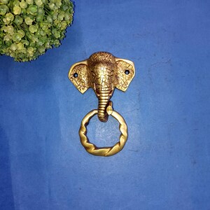 May include: A gold-colored brass door knocker shaped like an elephant's head with a ring pull.