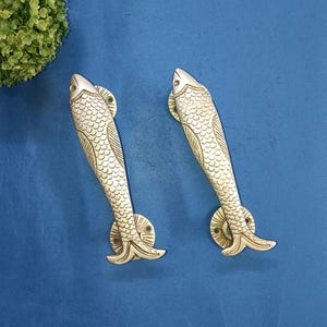 May include: Two silver fish-shaped door handles with detailed scales and a circular base. The handles are against a blue background.