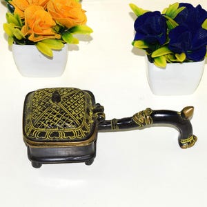 May include: An ornate, black and gold incense burner with a decorative handle. The square-shaped burner has a detailed, patterned design on the top and sides. Artificial flowers in white pots are in the background.