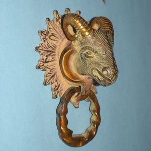 Peut inclure: Un heurtoir de porte en laiton en forme de tête de bélier avec des détails complexes. La tête de bélier est montée sur un motif floral décoratif.