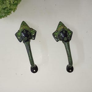 May include: Two black and green elephant-shaped drawer pulls. The elephant heads feature green detailing, and the handles have a textured design. These metal pulls are suitable for furniture or decorative use.