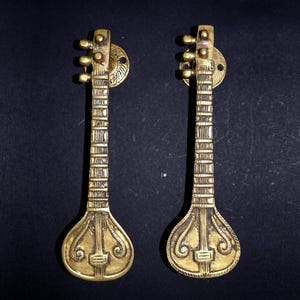 Puede incluir: Dos tiradores de puerta de color latón con forma de veena, un instrumento de cuerda indio. Cada tirador presenta un diseño detallado del cuerpo, el mástil y las clavijas del instrumento. Montados sobre placas circulares.