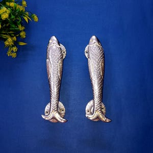 May include: A pair of silver-coloured, fish-shaped door handles. The handles have scale and fin details, set against a blue backdrop. These decorative handles are likely for a cabinet or door.
