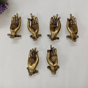 Puede incluir: Seis tiradores de puerta de metal dorado en forma de manos en posición de oración. Los tiradores tienen un diseño detallado y están montados sobre un fondo blanco.