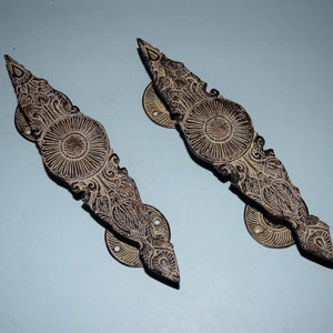 Op de afbeelding: Twee sierlijke messing deurgrepen met ingewikkelde bloemmotieven. De grepen zijn lichtbruin van kleur met een donkerbruine patina. Ze hebben een decoratieve ronde plaat met een centrale cirkel en een puntig uiteinde.