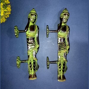 May include: Two bronze-colored door handles, each shaped like a standing Buddha figure. The handles have a dark bronze base with green accents, and a circular handle on one side. The Buddha figures are detailed with robes and facial features.
