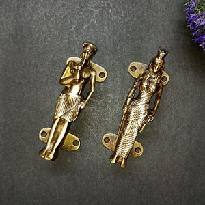 Op de afbeelding: Twee gouden metalen deurgrepen met decoratieve figuren. De grepen zijn gevormd als een man en een vrouw, beide met ingewikkelde details en een vintage stijl.