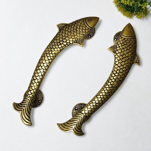 May include: Two brass-colored door handles shaped like fish. The handles have a textured scale pattern and are curved. The fish heads are at the top, and the tails are at the bottom. The handles are mounted on circular plates.
