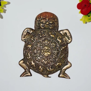 May include: A bronze-colored, turtle-shaped decorative item with an ornate, circular design on its shell. The turtle has a detailed face and legs. The shell features zodiac symbols and other intricate patterns. The item is likely a wall hanging or decorative piece.