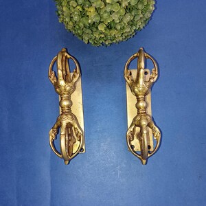 May include: A pair of gold-coloured metal door pulls with detailed patterns. Each pull has a central decorative feature with a handle, mounted on a rectangular plate. The background is a solid blue.
