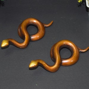 May include: Two gold-coloured snake sculptures with a curled tail and a gold-coloured head. The snakes are on a black surface.