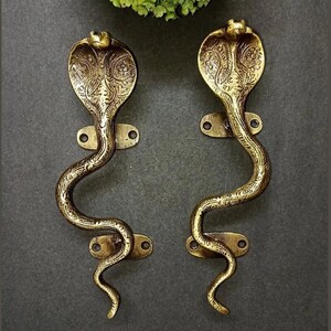 May include: A pair of brass-coloured door handles shaped like cobras. The handles feature detailed, engraved patterns and are mounted on rectangular plates. The cobra heads are flared, and the bodies curve. Decorative hardware.
