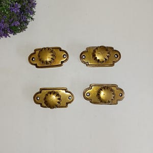 Puede incluir: Cuatro tiradores de cajón antiguos de latón. Cada tirador tiene una placa trasera rectangular con extremos redondeados y un pomo decorativo en forma de flor en el centro. Los tiradores son de un color latón cálido y envejecido, lo que sugiere encanto vintage.