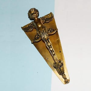 May include: A brass-toned door handle featuring a sculpted angel figure with outstretched wings. The handle is mounted on a triangular backplate with decorative detailing. The design incorporates floral and foliate elements.