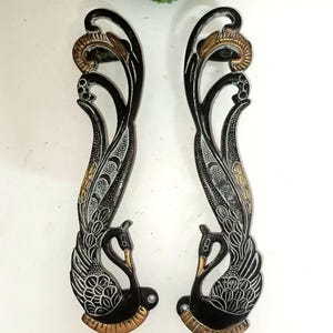 Puede incluir: Par de manijas de puerta decorativas negras con diseño de cisne. Las manijas presentan detalles intrincados con acentos dorados y plateados. El cuerpo y las plumas del cisne están detallados en plata, mientras que la parte superior e inferior tienen detalles dorados.