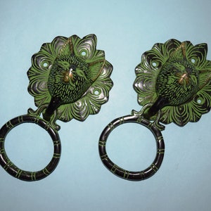 May include: Two green metal door knockers with a peacock design. Each knocker has a ring attached to the bottom.