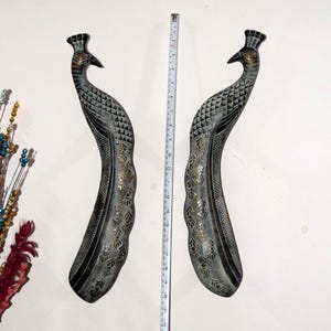 May include: Two decorative peacock sculptures. The sculptures are dark grey with intricate etched patterns and gold accents. Each peacock has a curved body and a detailed head with a crown. The sculptures are approximately 76 cm tall.
