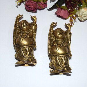 以下が含まれることがあります： 笑う仏陀の金色の金属製置物2つ。それぞれ腕を上げて笑顔をしています。置物は白い表面の上に立っています。