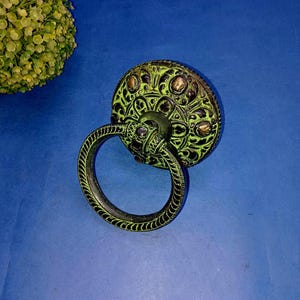 Könnte beinhalten: Ein runder Schubladengriff im antiken Stil mit dekorativem Design. Der Griff hat einen kreisförmigen Griff und eine detaillierte Oberfläche mit grünem und schwarzem Finish. Die Hardware befindet sich vor einem blauen Hintergrund.