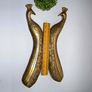 Puede incluir: Dos manijas de puerta doradas con forma de pavo real. Las manijas son de metal y presentan patrones de plumas detallados. Una regla amarilla se coloca entre las manijas. Las manijas miden aproximadamente 30 cm de largo.