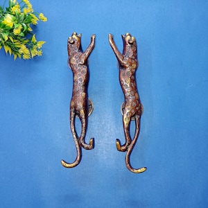 Könnte beinhalten: Zwei türgriffe in Form eines Leoparden in Bronzefarbe. Die Griffe sind mit Flecken versehen und stehen mit erhobenen Armen da. Die Griffe sind auf einem blauen Hintergrund montiert. Dekorative gelbe Blumen befinden sich in der oberen linken Ecke.