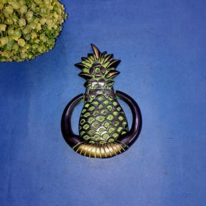 May include: A brass pineapple door knocker with a green patina finish. The knocker is in the shape of a pineapple with a ring for knocking.