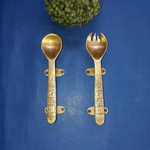 May include: A set of two gold-toned metal utensils, a spoon and a fork, with intricate designs on the handles. The utensils are mounted on a blue background.