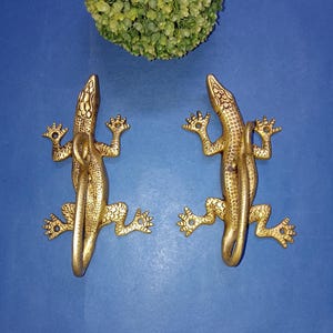 Könnte beinhalten: Zwei goldfarbene Metallhaken in Form von Eidechsen. Die Eidechsen sind nach rechts gerichtet und haben detaillierte Schuppen und eingerollte Schwänze.