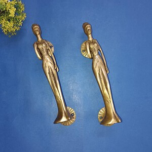 Peut inclure: Deux poignées de porte en laiton, chacune en forme de femme élancée dans une robe fluide. Les poignées sont de couleur or chaud et présentent des détails complexes. Les poignées sont montées sur une surface bleue.