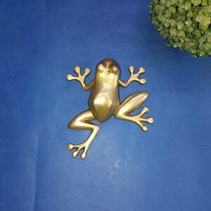 May include: A golden frog figurine with outstretched limbs. The frog is made of a shiny, metallic material and is positioned against a blue background. The frog's design is stylized, with a smooth, geometric body and detailed feet.