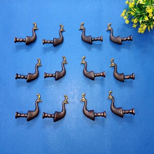 Könnte beinhalten: Elf bronzefarbene Schubladengriffe in Pfauform mit goldenen Akzenten werden auf einer blauen Oberfläche präsentiert. Jeder Griff weist ein detailliertes Pfauenmuster und eine kleine, runde Basis zur Montage auf.